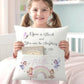 A young girl with pigtails holds a personalized white pillow featuring the quote 'Open a Book and You Can Be Anything To Be' and a pocket with a printed book and fairy illustrations. The scene is set in a soft, pastel bedroom environment.