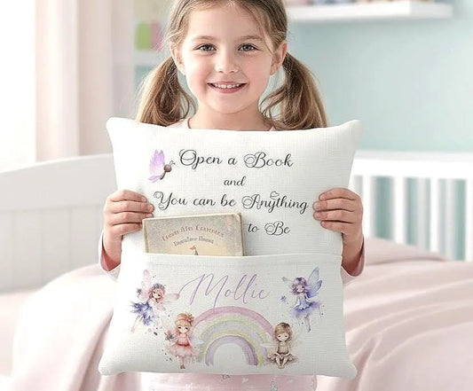 A young girl with pigtails holds a personalized white pillow featuring the quote 'Open a Book and You Can Be Anything To Be' and a pocket with a printed book and fairy illustrations. The scene is set in a soft, pastel bedroom environment.