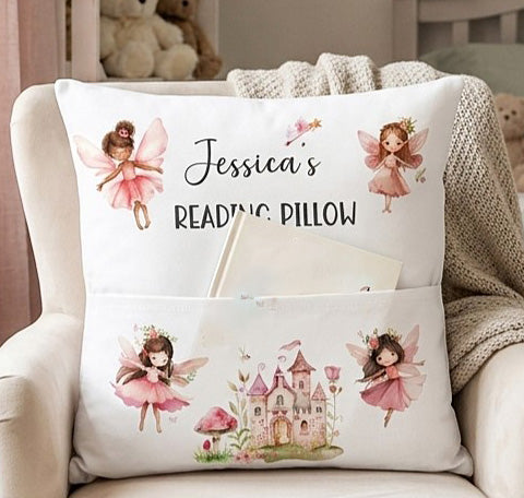 A personalized white reading pillow with fairy and castle illustrations, resting on a beige armchair with a knitted blanket. The pillow features the text 'Jessica's READING PILLOW' and includes a front pocket holding a book.