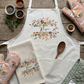 A styled flat-lay of a white kitchen apron with a floral 'Rosie' design, accompanied by matching floral oven mitts, a waffle-knit tea towel, and quilted oven mitts on a rustic wooden background.