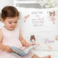 A toddler in a pale pink dress sits on a white rug, reading a book in front of a decorative white pillow. The pillow features fairy illustrations and the printed phrase 'EVERY TIME YOU open a book A LITTLE MAGIC falls out' plus the name 'Olivia' in script.