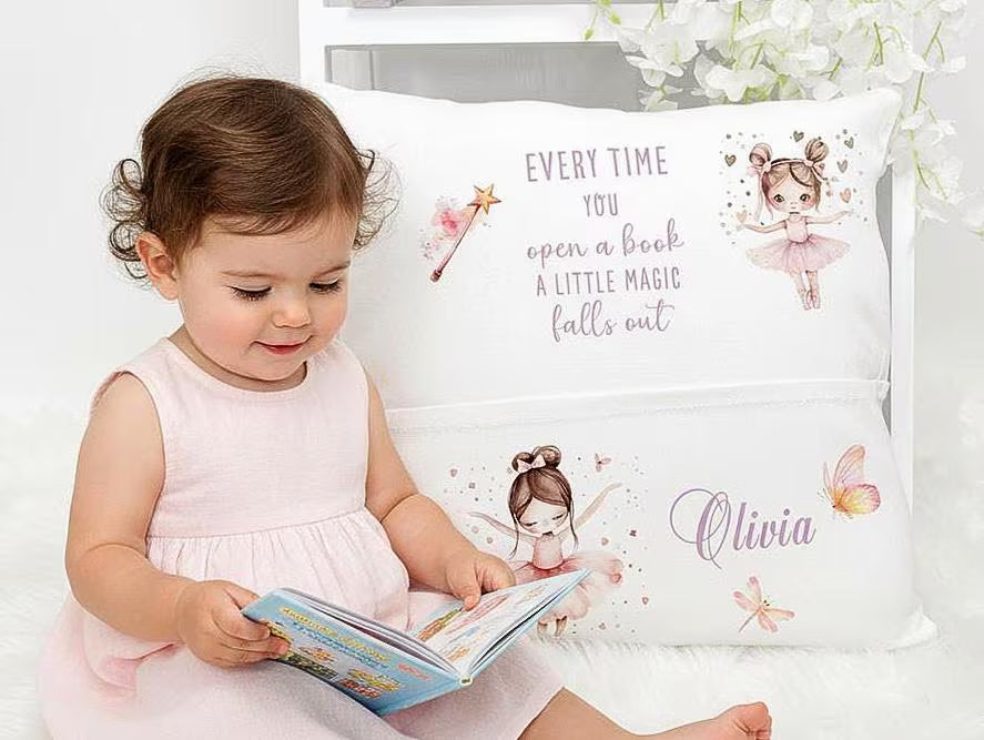 A toddler in a pale pink dress sits on a white rug, reading a book in front of a decorative white pillow. The pillow features fairy illustrations and the printed phrase 'EVERY TIME YOU open a book A LITTLE MAGIC falls out' plus the name 'Olivia' in script.