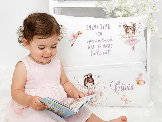 A toddler in a pale pink dress sits on a white rug, reading a book in front of a decorative white pillow. The pillow features fairy illustrations and the printed phrase 'EVERY TIME YOU open a book A LITTLE MAGIC falls out' plus the name 'Olivia' in script.