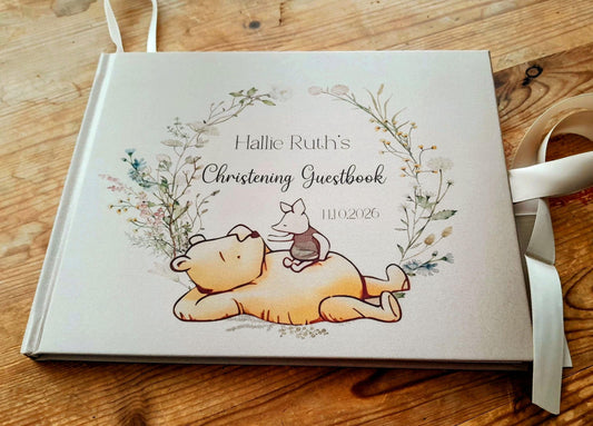 A personalized baby christening guestbook featuring Winnie the Pooh and Tigger, resting on a rustic wooden table with a white ribbon tied to its spine.
