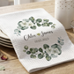 White towel with green leaf design and personalized text on a wooden table.
