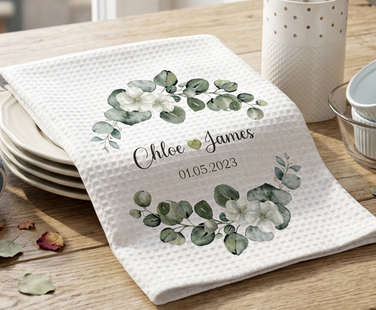 White towel with green leaf design and personalized text on a wooden table.