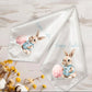 Two white satin Easter-themed napkins with printed illustrations of a bunny in a blue outfit holding a pink egg, displayed on a light wood background with decorative flowers.