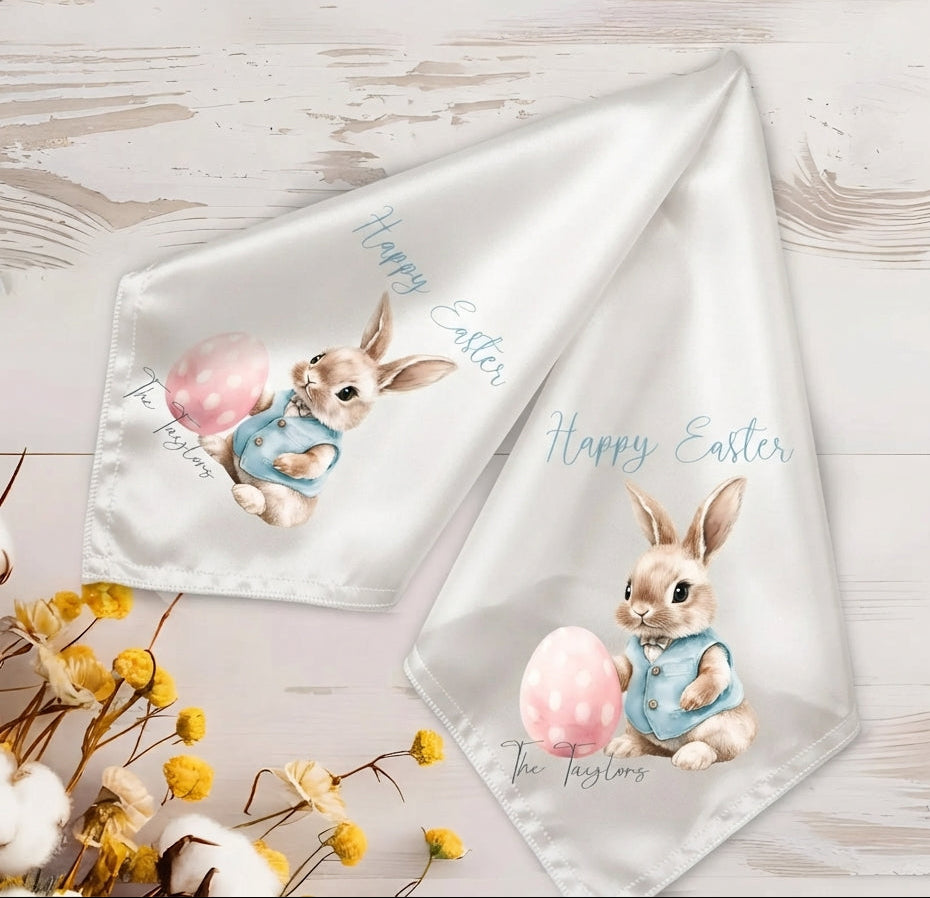 Two white satin Easter-themed napkins with printed illustrations of a bunny in a blue outfit holding a pink egg, displayed on a light wood background with decorative flowers.