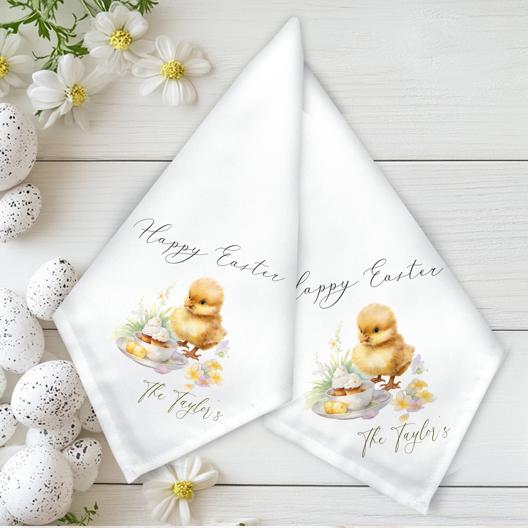 A set of two white fabric napkins with a personalized floral Easter chick design, placed on a table with decorative Easter eggs and flowers in the background.