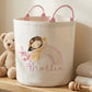 A cream fabric storage bin with pink handles, decorated with a cartoon girl, a pastel rainbow, and the name 'Mollie' in script. The bin sits on a wooden shelf next to a teddy bear, folded blankets, and wooden toys in a softly lit nursery setting.