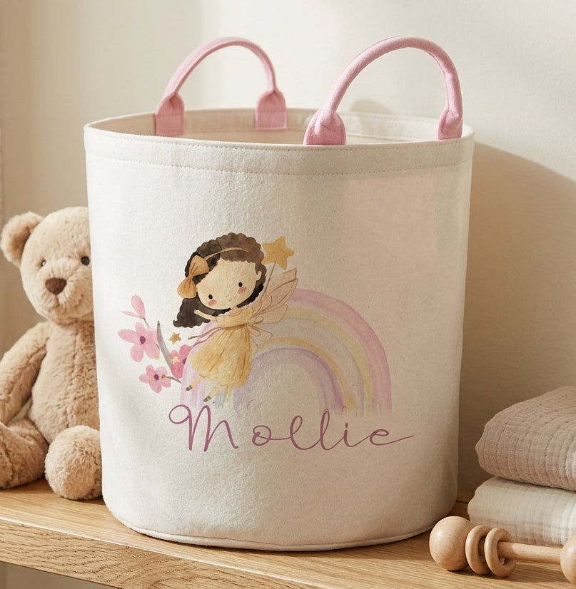 A cream fabric storage bin with pink handles, decorated with a cartoon girl, a pastel rainbow, and the name 'Mollie' in script. The bin sits on a wooden shelf next to a teddy bear, folded blankets, and wooden toys in a softly lit nursery setting.