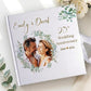 A personalized hardcover wedding anniversary photo book with a white cover, featuring a printed couple's portrait framed by green foliage, gold script text reading 'Emily & David,' and '25th Wedding Anniversary 2001 ♥ 2026.' The book is styled on a light surface with white flowers and green leaves around it.