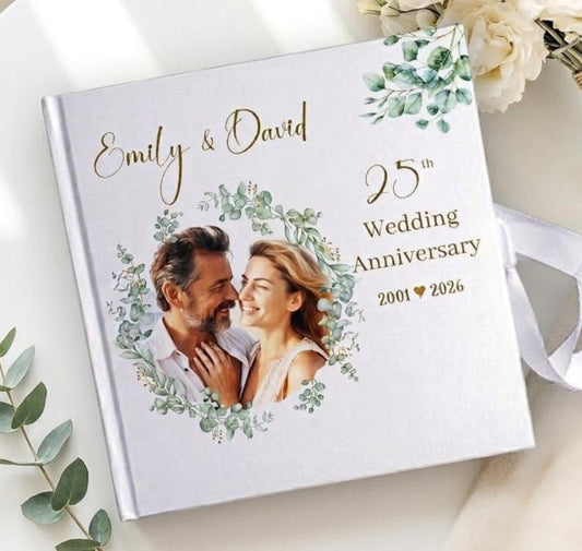 A personalized hardcover wedding anniversary photo book with a white cover, featuring a printed couple's portrait framed by green foliage, gold script text reading 'Emily & David,' and '25th Wedding Anniversary 2001 ♥ 2026.' The book is styled on a light surface with white flowers and green leaves around it.