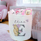 A personalized fabric storage basket in a nursery setting, featuring the name 'Eliza' and a large letter 'E' with a cartoon dog illustration. The basket is filled with plush teddy bears and is positioned next to a white crib.