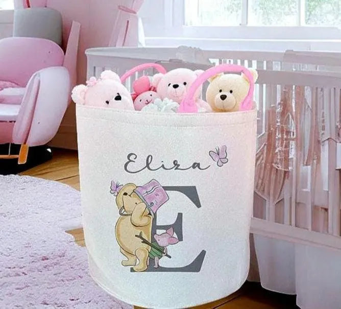 A personalized fabric storage basket in a nursery setting, featuring the name 'Eliza' and a large letter 'E' with a cartoon dog illustration. The basket is filled with plush teddy bears and is positioned next to a white crib.