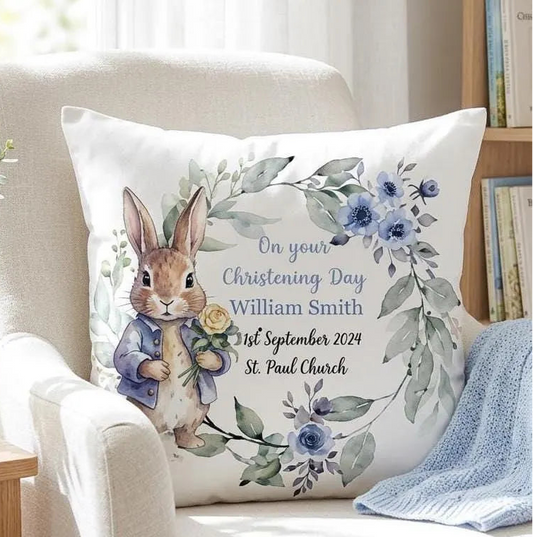A personalized square white pillow featuring a watercolor illustration of Peter Rabbit in a blue jacket holding a yellow rose, surrounded by blue flowers and green leaves. The text reads 'On your Christening Day William Smith 1st September 2024 St.