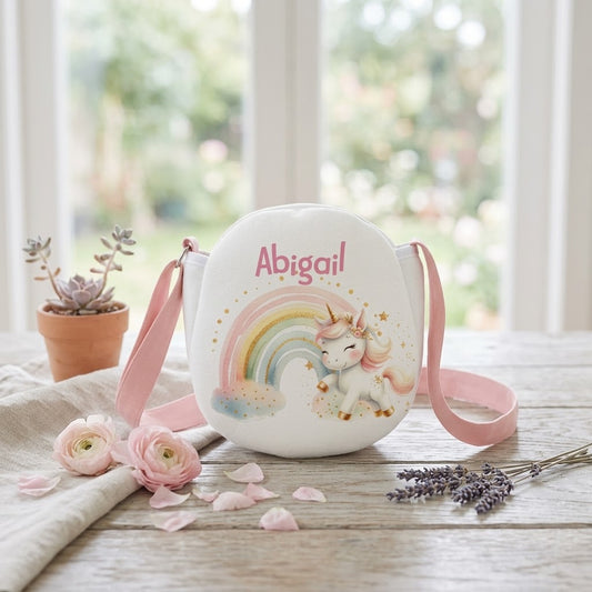 A personalized white crossbody bag with a pink strap, featuring a printed unicorn and rainbow design with the name 'Abigail' in pink script. The bag is styled on a rustic wooden table with pink flowers, lavender, and a small potted succulent in front of a bright window.