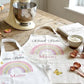 Two personalized white kitchen aprons laid out on a rustic wooden table, featuring pastel rainbow designs and the names 'Star Baker' and 'Mummy' with 'Mollie' below. A cream stand mixer, baking ingredients, measuring cups, and a book are arranged around them in a bright kitchen setting.