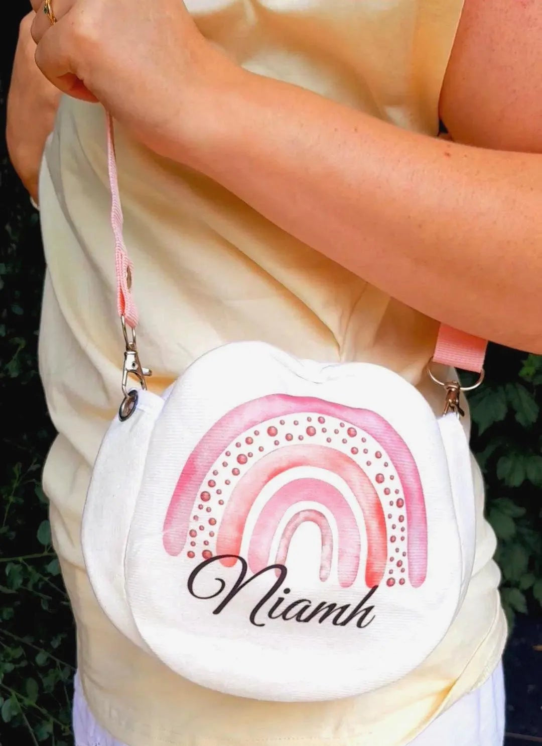 A person wearing a cream top and white skirt carries a small white crossbody bag with a pink watercolor rainbow design and the name 'Niamh' in black script.