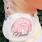 A cream-colored crossbody bag with a pink watercolor rainbow design and the name 'Niamh' printed in black script. The bag is shown being worn outdoors by a person wearing a pale yellow top and white skirt.