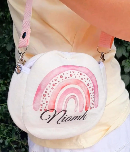 A cream-colored crossbody bag with a pink watercolor rainbow design and the name 'Niamh' printed in black script. The bag is shown being worn outdoors by a person wearing a pale yellow top and white skirt.