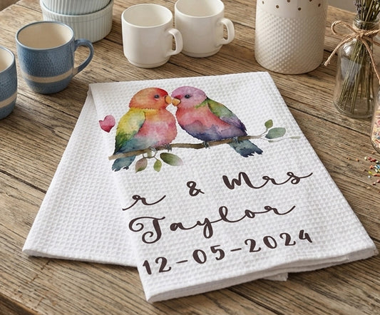 Personalized kitchen towel with colorful birds and text on a wooden table with mugs.