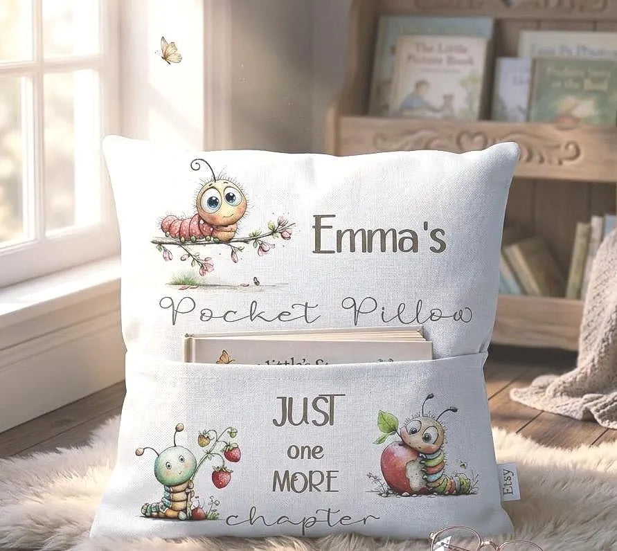 A personalized pocket pillow displayed in a cozy room setting. The white pillow features printed cartoon illustrations of a pink caterpillar and a green ladybug with the text 'Emma's Pocket Pillow' and 'JUST ONE MORE chapter' in a whimsical font.